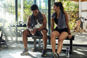 Two people rehydrating with water bottles post workout in the gym, sitting on a bench with towels.