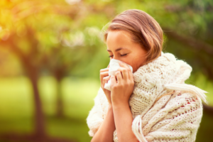 Woman sneezing due to season change sickness symptoms, wrapped in a cozy scarf outdoors during fall.